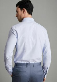 Lichtblauw, overhemd met lange mouwen en een klassieke kraag, voorzien van knoopmanchetten. Gekleed met grijze op maat gemaakte broek, die een slim fit laat zien.