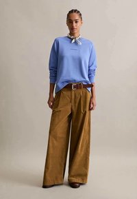 Mulher de pé, vestindo um sweatshirt azul claro, calças largas bege com um cinto castanho, sapatos castanhos e um lenço branco no pescoço, fundo neutro.