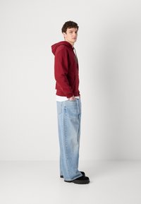 Felpa cappucciata bordeaux con tasca frontale, abbinata a jeans di denim baggy azzurri chiari e zoccoli neri, mostrata di lato.
