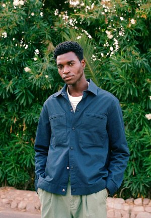 Ung mand med hænderne i lommerne iført en marineblå jakke og lysegrønne bukser står foran grønne bladplanter med hvide blomster.