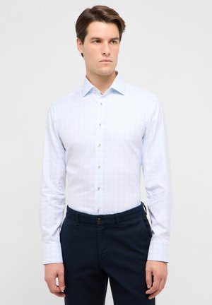 Giovane uomo con indosso una camicia celeste a bottoni con un motivo a quadretti sottile e pantaloni blu scuro, in piedi davanti a uno sfondo semplice.