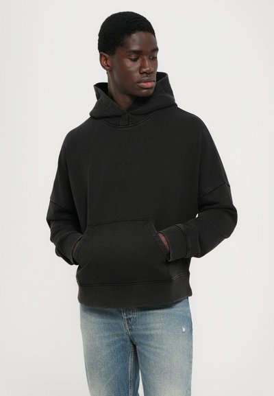 Schwarzer Kapuzenpullover mit einer Fronttasche und gerippten Bündchen. Besitzt ein entsprechendes Logo auf der Brust und eine lässige Passform. Getragen mit hellblauen Jeans.