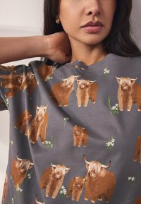 Grå stoftop med mønster af brune højlandskvæg og kalve med grønt græs og hvide blomster. Korte ærmer og afslappet pasform.