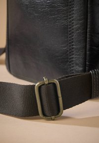 Black leather bag corner with a textured finish, featuring a brown woven strap secured by a rectangular antique bronze buckle.
