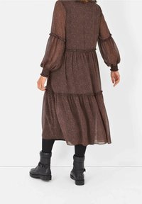 Mujer con un vestido midi de leopardo de manga larga marrón con falda de capas, leggings negros y botas negras de media pantorrilla con hebilla, de pie de espaldas.
