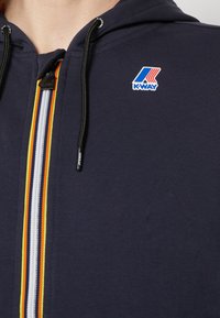 Námořnická mikina na zip z měkké látky s vertikálním proužkovaným vzorem v oranžové, bílé a žluté barvě, s logem K-Way na hrudi.