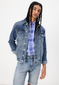 Denim-takki eri sinisissä sävyissä, joissa on klassiset taskut, valkoiset napit ja käärityt hihat, käytetty sinisen ruudullisen paidan ja farkkushortsien päällä.