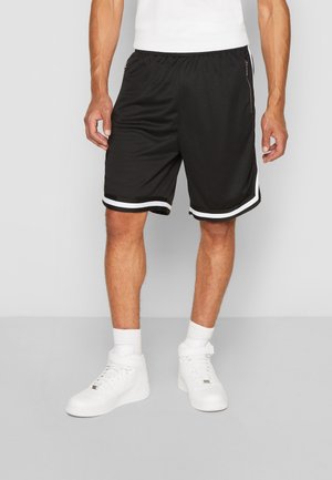 Homme portant un short de sport noir avec un détail de bande blanche, des chaussettes blanches et des baskets blanches montantes, debout sur un sol gris.