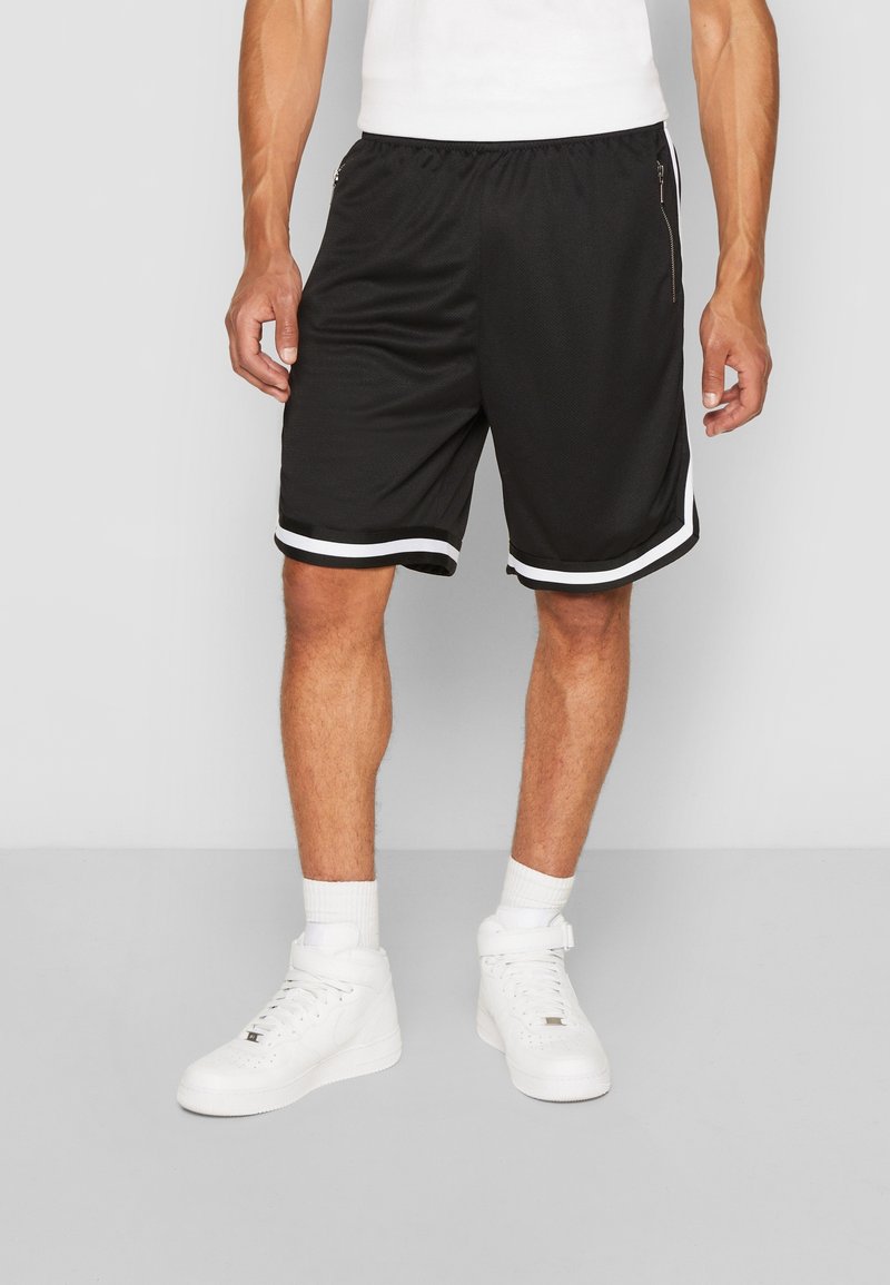 Homme portant un short de sport noir avec un détail de bande blanche, des chaussettes blanches et des baskets blanches montantes, debout sur un sol gris.