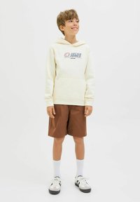 Haut à capuche crème avec logo, poche avant ; shorts marron, longueur genou. Chaussettes blanches avec baskets à rayures noires. Tenue simple et décontractée.
