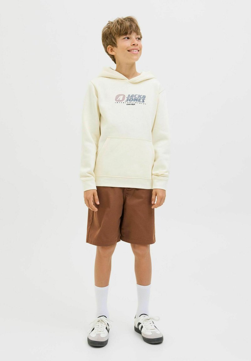 Haut à capuche crème avec logo, poche avant ; shorts marron, longueur genou. Chaussettes blanches avec baskets à rayures noires. Tenue simple et décontractée.