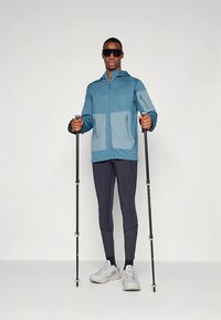 Könnyű kék dzseki sötétebb részletekkel, cipzáras zsebekkel, fekete leggingshez és szürke túracipőhöz párosítva; túrabotokkal a kezében.