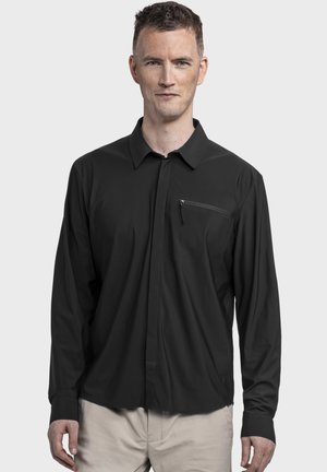 Mann trägt ein schwarzes, langärmliges Hemd mit Knöpfen und einer Reißverschlusstasche auf der Brust sowie beige Hosen, steht vor einem einfarbigen Hintergrund.