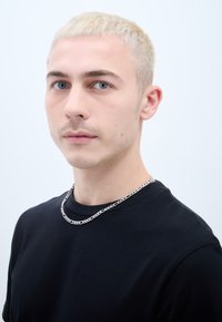 Juodas medvilninis marškinėliai su apykakle, dėvimi su sidabrine grandinėle. Modelis turi trumpus platininės blondinės plaukus ir mėlynas akis.