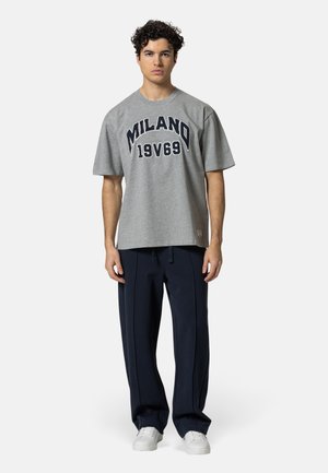 Homme portant un t-shirt gris "MILANO 19V69", un pantalon large bleu marine et des baskets blanches, debout face à la caméra sur un fond uni.