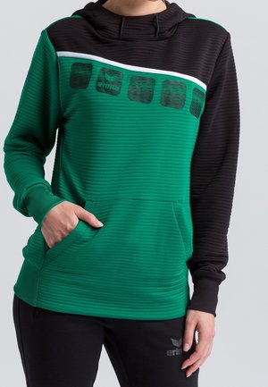 Osoba ubrana w zielono-czarną prążkowaną bluzę z kapturem i przednią kieszenią oraz czarne spodnie, ręka w kieszeni, stojąca na jednolitym tle.