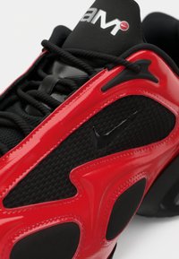 Zapatilla deportiva roja y negra con una malla negra texturizada, superposiciones rojas brillantes y cordones negros. Presenta un pequeño logo negro de Nike en el lateral.