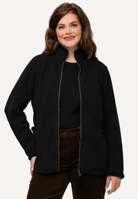 Černá fleecová bunda s vysokým límcem, zipem vpředu a bočními kapsami. Měkká textura, strukturovaný design. Nosit s tmavě hnědými kalhotami.