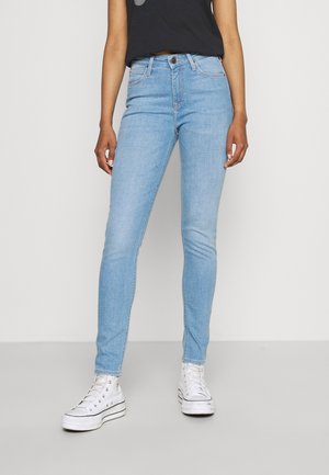 Jeans ajustados de mezclilla azul claro con una cintura de altura media, que cuentan con un cierre de botón, bolsillos clásicos y una textura suave. Combinados con zapatillas blancas.