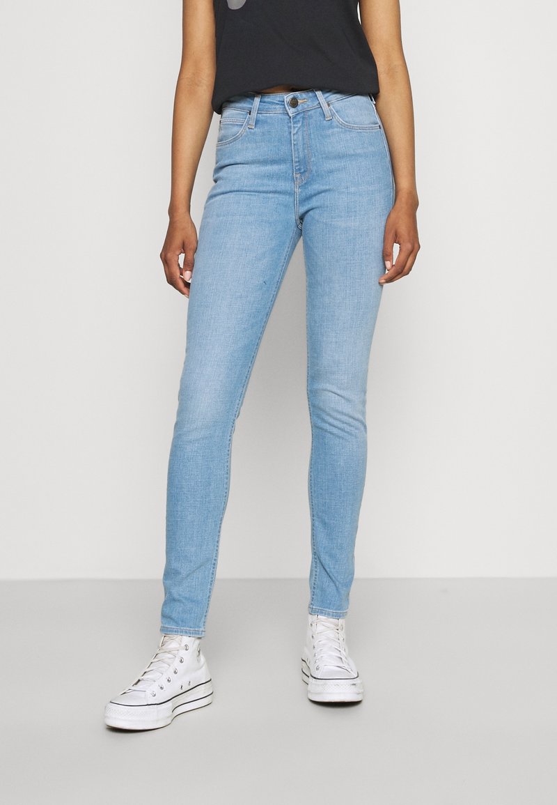 Jeans ajustados de mezclilla azul claro con una cintura de altura media, que cuentan con un cierre de botón, bolsillos clásicos y una textura suave. Combinados con zapatillas blancas.