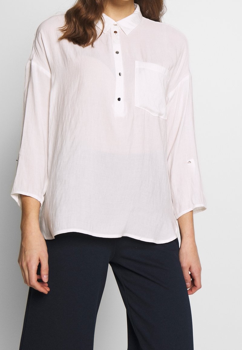 Chemise rose clair à manches longues avec un devant boutonné, une poche sur le côté gauche et des poignets retroussés, fabriquée en tissu texturé et léger.