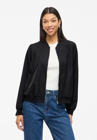 Γυναίκα που χαμογελά, φορώντας ασημένια κρεμαστά σκουλαρίκια, μαύρο φερμουάρ πανωφόρι πάνω από άσπρο τοπ, και ψηλόμεσα μπλε τζιν, με φόντο έναν απλό τοίχο.