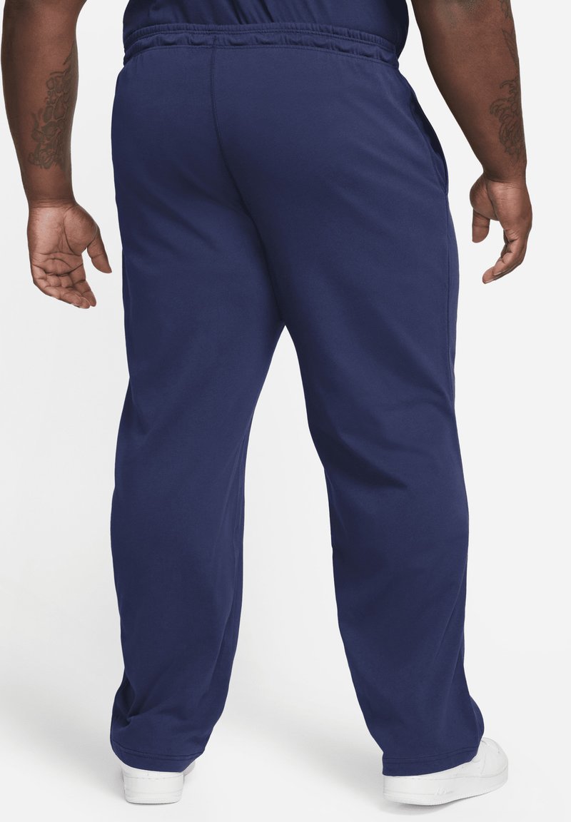 Pantalon de survêtement bleu marine avec une taille élastique et des poches latérales. Le matériau semble lisse et léger. Baskets blanches visibles en bas.