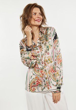 Kvinde smiler iført en langærmet skjorte med et farverigt blomstret og paisley mønster og hvide bukser mod en ensfarvet baggrund.