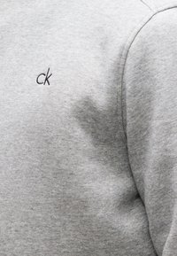 Κοντινό πλάνο ανοιχτογκρι υφάσματος με μικρό μαύρο κεντημένο λογότυπο "ck" στην περιοχή του στήθους μιας φούτερ μπλούζας.