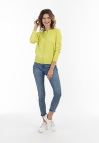 Cardigan giallo brillante con colletto rotondo e chiusura con bottoni, abbinato a jeans blu attillati e sneakers bianche. Texture liscia, stile casual.
