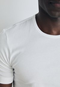 T-shirt en coton blanc à col rond, avec des manches courtes, texture lisse et design ajusté, près du cou et de l'épaule.