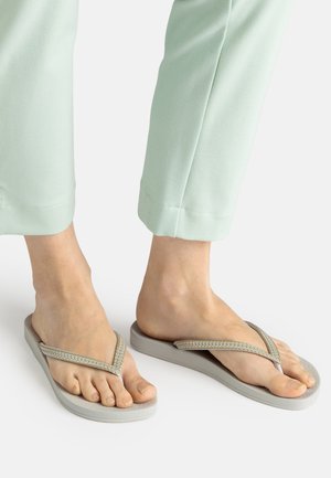 Füße in hellgrauen Flip-Flops mit dekorativen Riemen, kombiniert mit mintgrünen Cropped-Hosen, stehend auf einer weißen Oberfläche.