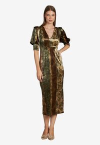 Donna in piedi che indossa un vestito midi di velluto lucido con maniche a sbuffo nei toni del marrone e del verde, abbinato a tacchi beige con dettagli decorativi.