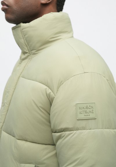 Jasnozielona kurtka puchowa z wysokim kołnierzem, posiadająca pikowaną teksturę oraz prostokątną naszywkę z logo Maison Kitsuné na ramieniu.