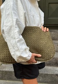 Sac à main tressé en olive avec une forme ovale et une surface texturée, tenu sous le bras par une personne portant une veste blanche et un short noir.