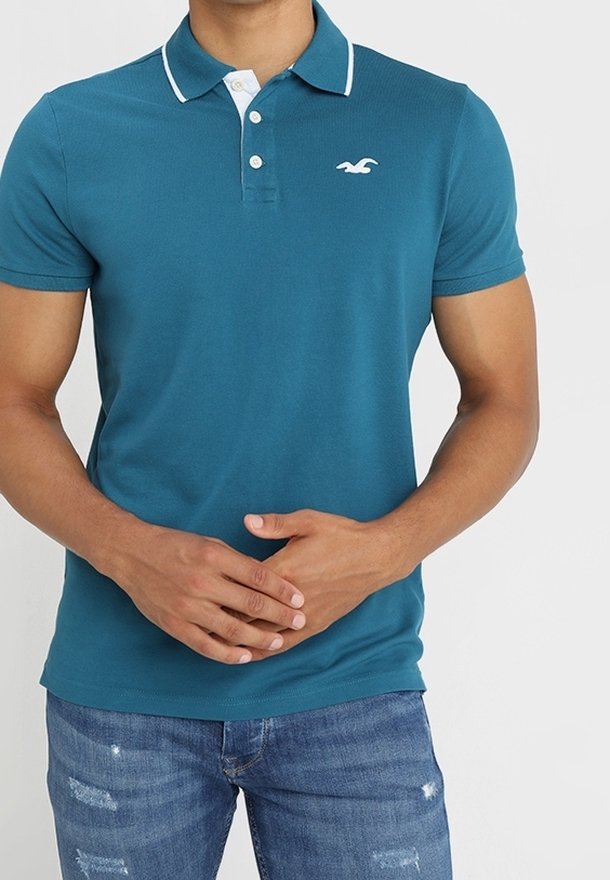 Teal polo paita, joka on valmistettu sileästä kankaasta. Siinä on valkoinen kauluksen reuna, kolmen napin hakaset ja pieni logo vasemmassa rinnassa.