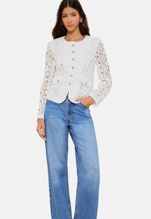 Vrouw draagt een witte kanten jas met gouden knopen en blauwe hoog getailleerde wijde jeans, staand met één hand in de zak.