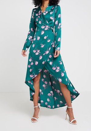 Kobieta w turkusowej sukience kopertowej w różowy kwiatowy wzór z asymetrycznym dołem, połączonej z białymi szpilkami na pasku wokół kostki, stojąca na jednolitym tle.