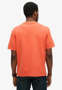 Mand iført en ensfarvet orange kortærmet t-shirt og mørkeblå jeans, vist bagfra mod en hvid baggrund.