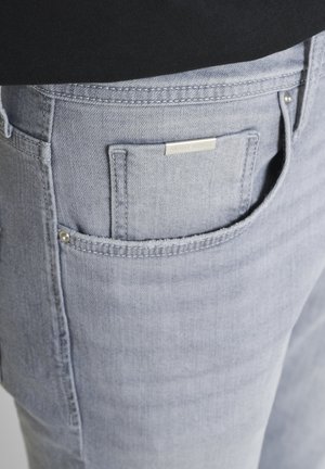 Hellgraue Denim-Jeans mit Taschen-Detail, Metallmarkenetikett und Nieten, kombiniert mit einem schwarzen Oberteil.