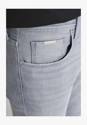 Detalj džepova svijetlosivih teksas hlača s metalnom oznakom marke i zakovicama, nošene s crnim gornjim dijelom.