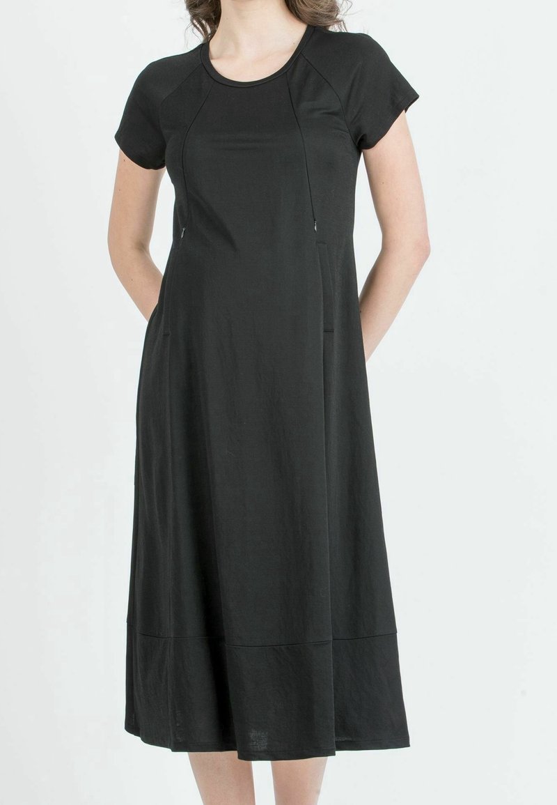 Femme portant une robe midi noire unie à manches courtes, col rond et coupe décontractée, debout les mains derrière le dos.