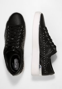 Černé kožené tenisky s bílými podrážkami, s kovovými hroty po stranách a lesklým akcentem na patě. Černé tkaničky a značení uvnitř.