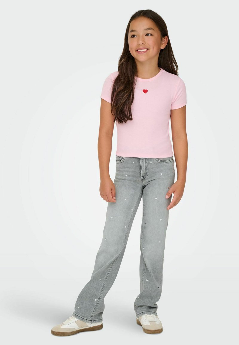 Dievča s dlhými tmavými vlasmi, oblečené v ružovom tričku s krátkym rukávom s červeným srdcom a sivých džínsoch s malými bielymi srdiečkami, stojí a usmieva sa.