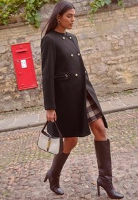 Cappotto di lana nero con bottoni dorati e spacco laterale, abbinato a una gonna a quadri. Stivali alti in pelle marrone con tacco e una borsa bicolore.