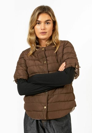 Frau mit blondem Haar, trägt eine braune gesteppte Jacke mit kurzen Ärmeln über einem langärmeligen schwarzen Oberteil und dunkelgraue Hose, Arme verschränkt.