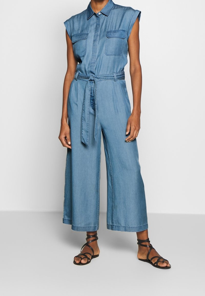 Tuta di denim in azzurro chiaro, con maniche corte, colletto e vita con cinturino. Include due tasche sul petto e gambe larghe.