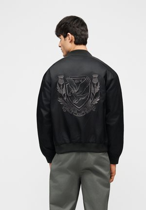 Uomo che indossa una giacca bomber nera con stemma di aquila ricamato, testo "1874" e "Lyle and Scott" sul retro, abbinata a pantaloni grigi.