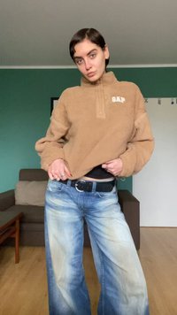 Άτομο με μπεζ φλις πουλόβερ και φαρδιά, ξεβαμμένα μπλε τζιν στέκεται μέσα σε κλειστό χώρο, κρατώντας το πουλόβερ λίγο σηκωμένο με το ένα χέρι.