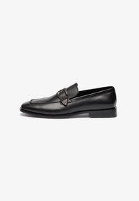 Mocasines de cuero negro con un diseño elegante, puntera puntiaguda y una hebilla decorativa en el lado lateral. Suela plana de goma y acabado pulido.